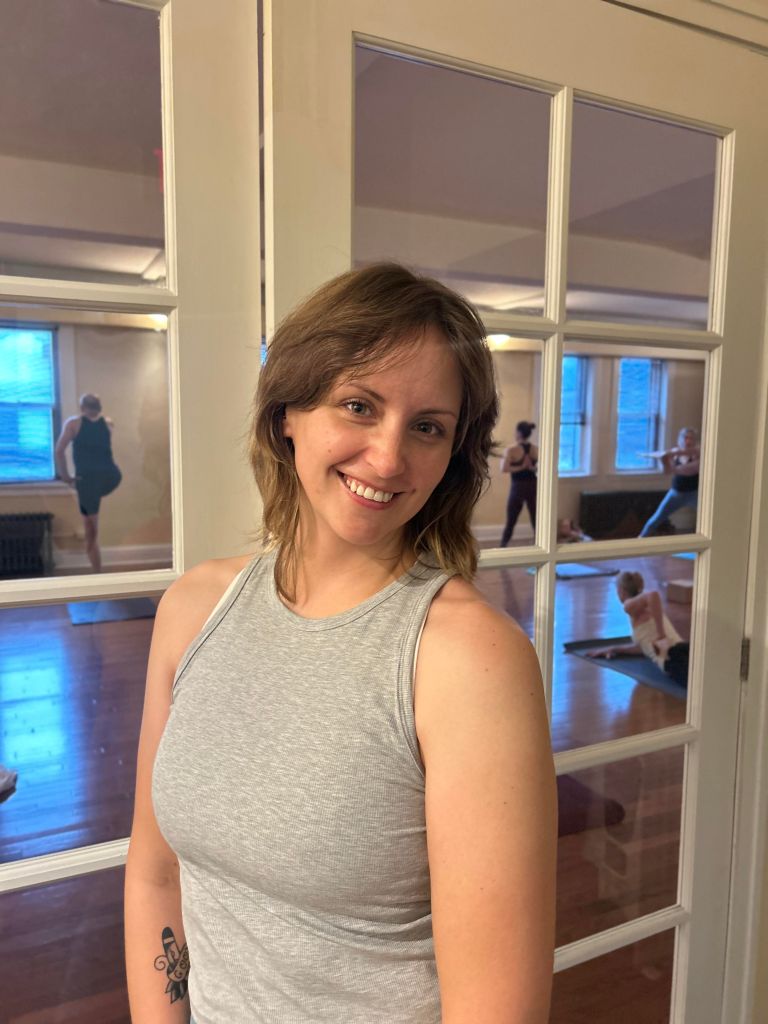 A smiling individual with shoulder-length hair stands in front of a glass-paneled door, showcasing a yoga studio in the background with practitioners engaged in yoga poses.